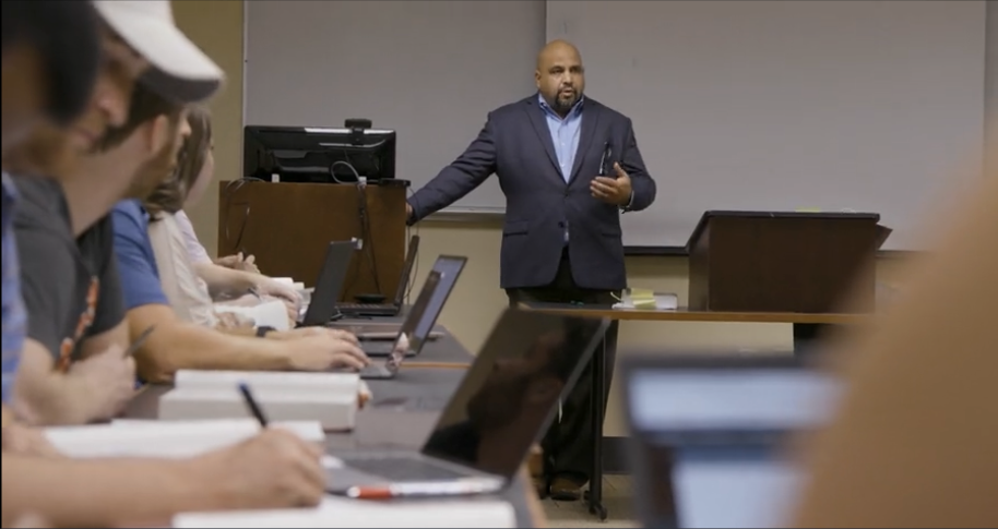 A professor stands behind a podium and addresses students in a classroom