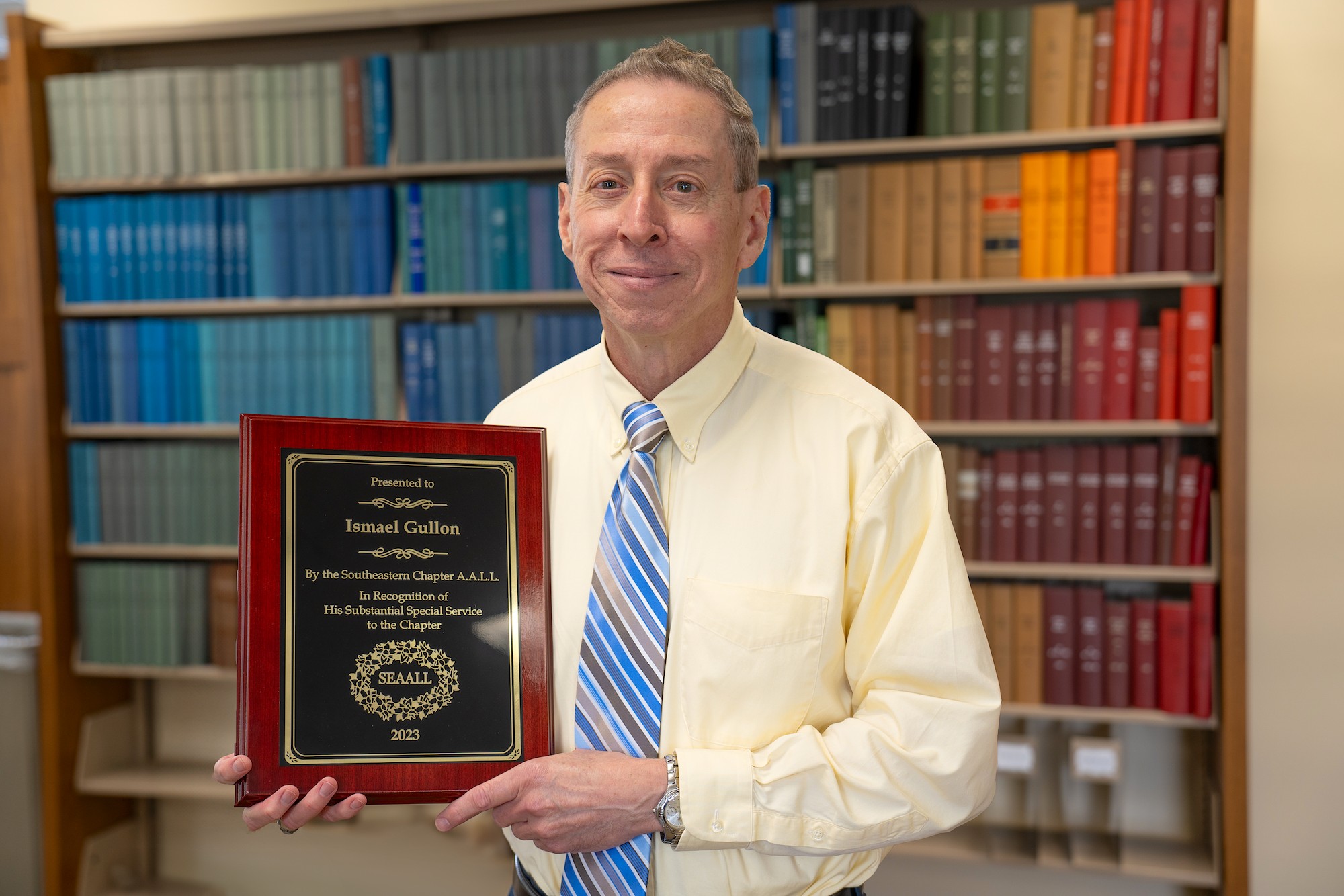 Ismael Gullon holds a plaque, standing in front of a wall of books at the Mercer Law library