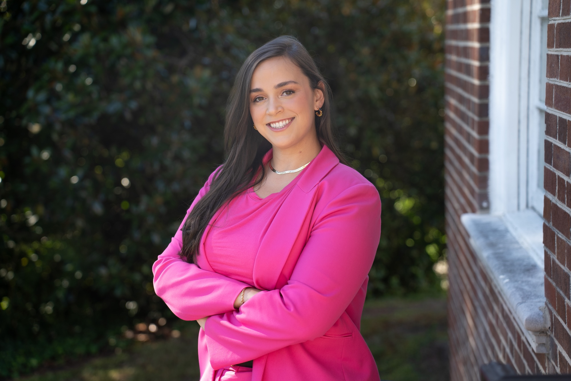 Headshot of Hannah Frantz in a pink suit standing in front of trees and beside a brick building