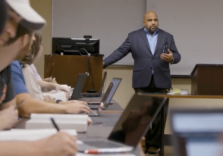 A professor stands behind a podium and addresses students in a classroom