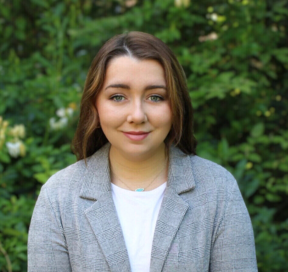 Olivia Brillinger in a gray jacket and white shirt in front of a green leafy background