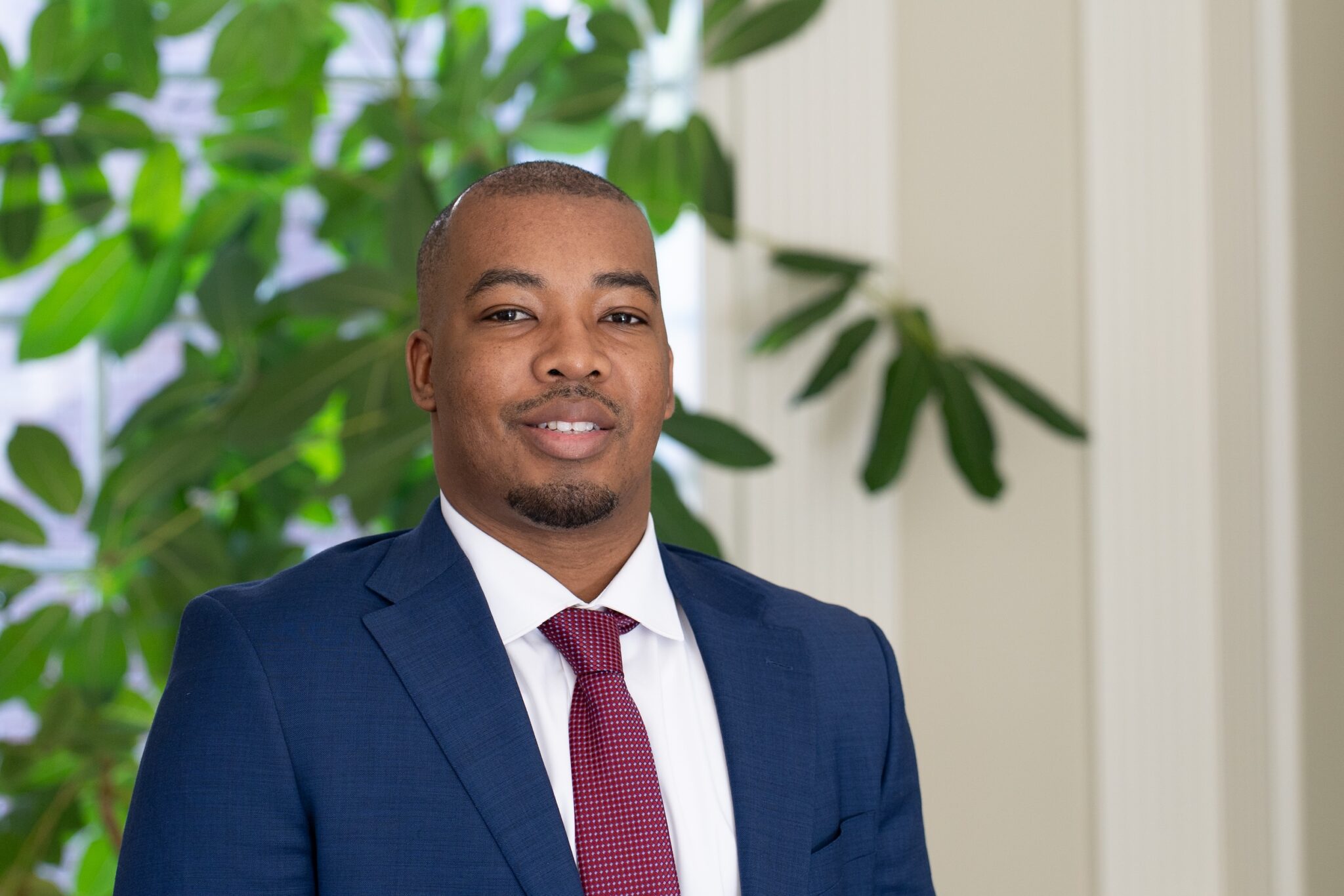 Lucious Moore in a blue jacket, white shirt, and red tie standing in front of a large green plant