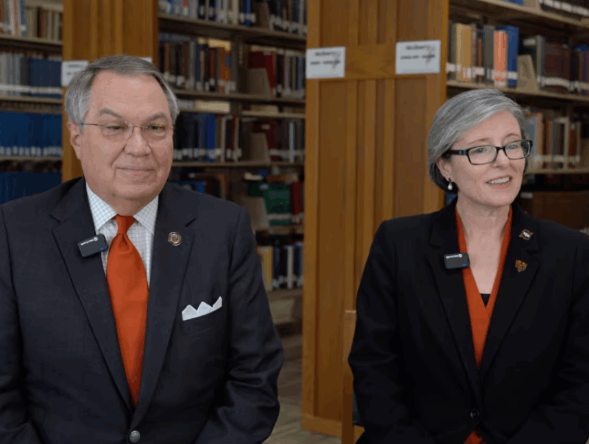 Image of a man in a black jacket and orange tie and a woman in a black jacket and an orange tie sitting in a library
