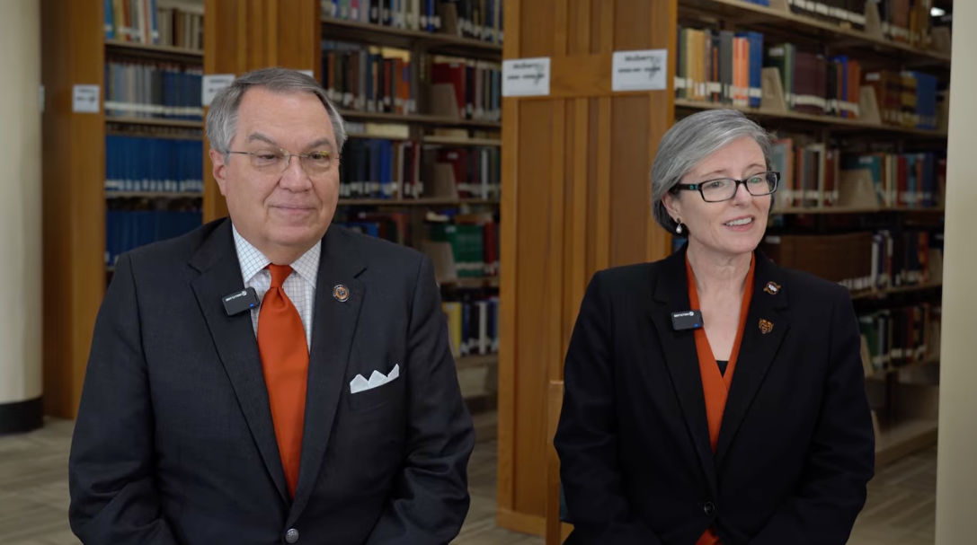 Image of a man in a black jacket and orange tie and a woman in a black jacket and an orange tie sitting in a library