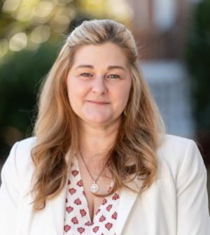 Woman in white jacket stands in front of greenery and a brick building