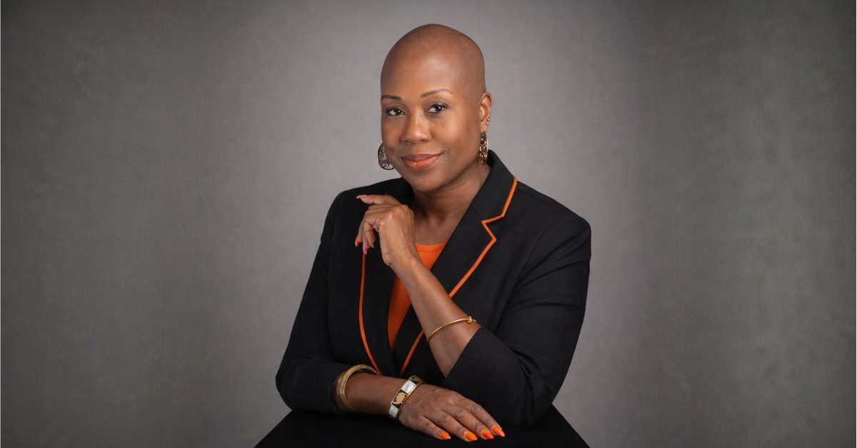 A woman sits in front of a gray background, wearing a black blazer and an orange shirt
