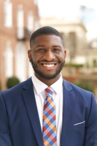 Shaun Neville stands outside in a blue jacket, white shirt, and orange and blue checkered tie