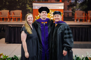 One woman in a black dress stands with two men in graduation regalia