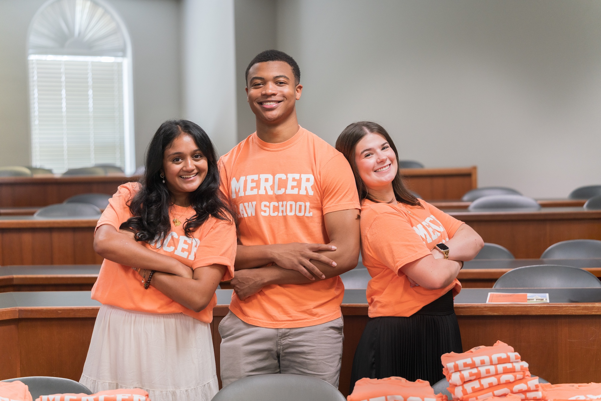 Two female students stand back-to-back with a male student, all wearing orange Mercer Law t-shirts
